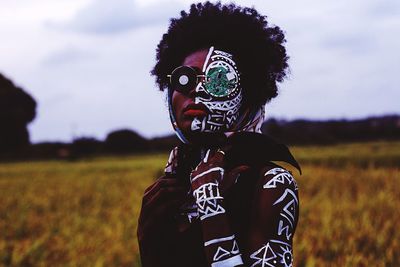 Portrait of young woman with body paint standing on field against sky