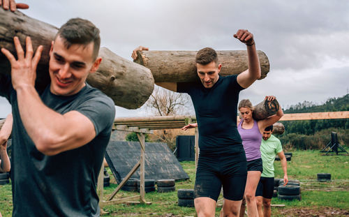 Friends carrying logs while standing on field against sky