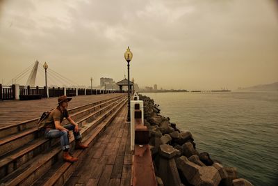 Man sitting on pier by sea against sky