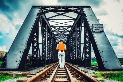 Rear view of man standing on railroad bridge against cloudy sky