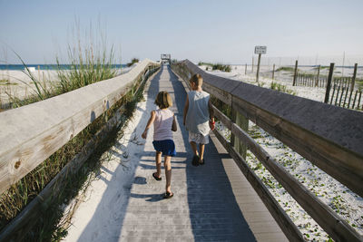 Rear view of siblings walking on boardwalk at beach against clear sky