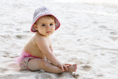 Cute little baby girl in pink swimming trunks and panama sitting on sand at beach.