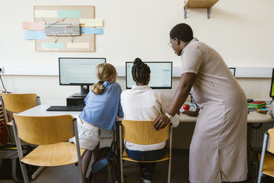 Side view of female teacher explaining girls using computer while sitting in classroom at school