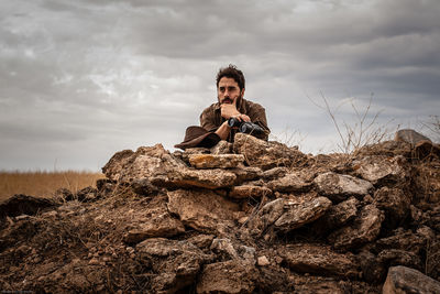 Thoughtful man holding binoculars standing behind rock formation against cloudy sky