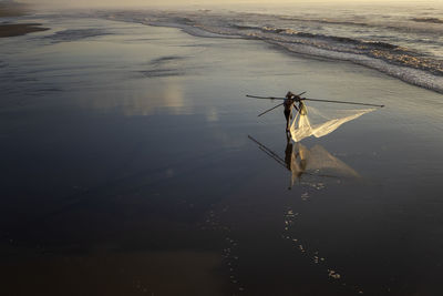 Fisherman with fishing nets standing at beach against sky during sunset