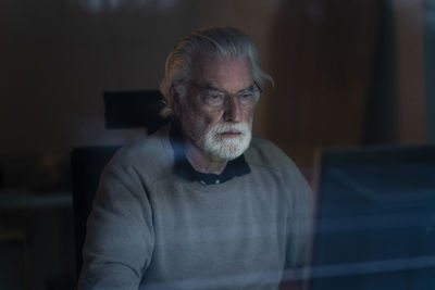 Senior man using computer at home during night seen through glass