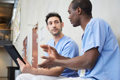 Male nurse gesturing while discussing over digital tablet with coworker in corridor at hospital