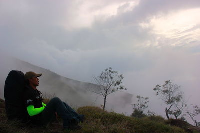 Side view of man looking away while sitting on mountain against sky