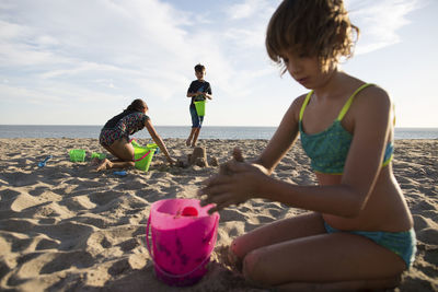 Siblings making sand castle at beach against sky