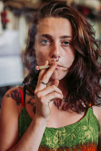 Young red haired female hippie in colorful dress smoking blunt in stylish room looking at camera