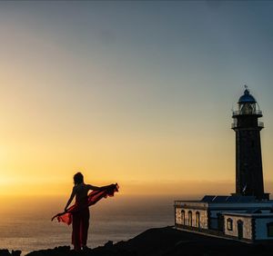 Silhouette woman standing on cliff by lighthouse against sea at sunset