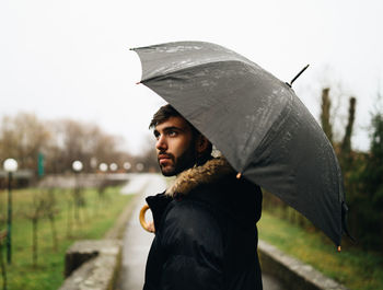 Side view of young man holding umbrella while standing outdoors during rainy season