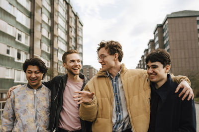 Smiling male friends talking while walking with arms around in city