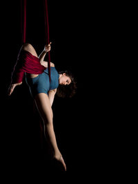 Young woman hanging on fabric while dancing against black background