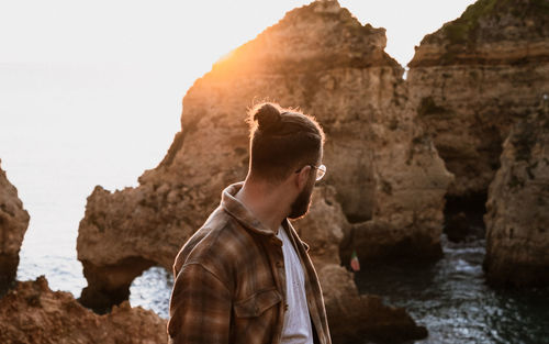 Young man looking at rock formation in sea with sunrise coming up