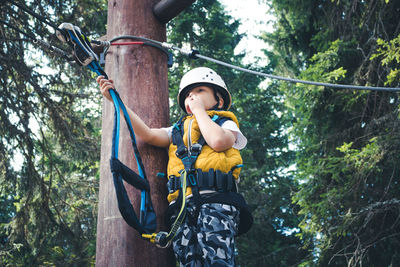 Low angle view of kid standing on treetop while having rope course at adventure park.