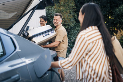 Rear view of woman charging car while happy man loading luggage with son in trunk