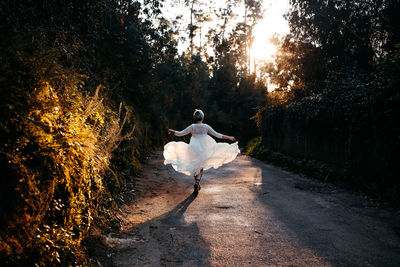 Full body back view of anonymous female wearing white dress walking on rural road among green trees in nature on evening time