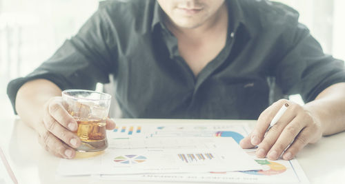 Midsection of businessman having drink and cigarette while reading graphs on table