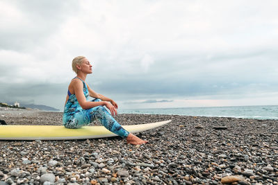 Young blond woman surfer sitting on surfboard looking to the sea on the beach in cloudy day.