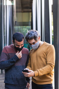 Man in mask sharing smartphone with male friend while standing on street in city