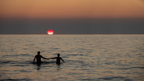 Silhouette men in sea against sky during sunset