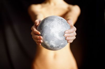 Midsection of shirtless woman holding moon against curtain