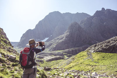 Rear view of hiker with backpack photographing mountains while standing on field 