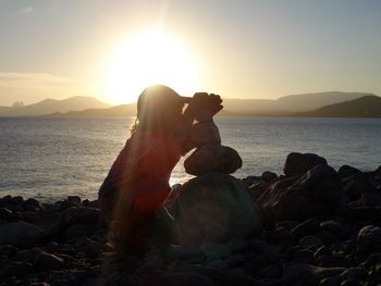 Boy stacking stones at beach against sky during sunset