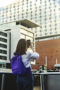 Woman with backpack photographing elbphilharmonie through smart phone, hafencity, hamburg, germany