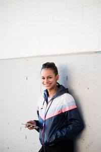 Portrait of smiling young woman listening to music while standing by wall