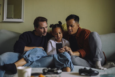 Girl using smart phone while sitting with gay parents on sofa at home