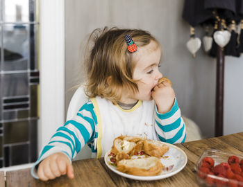 Cute girl eating bread while sitting by table at home