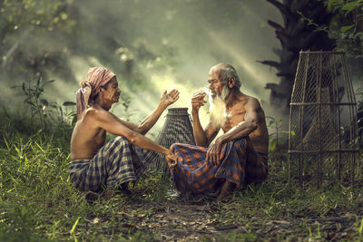 Shirtless man smoking cigarette while sitting with friend on field