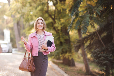 Stylish woman 45-50 year old holding books and magazines with leather bag outdoors.