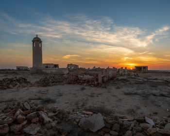 Lighthouse by sea and buildings against sky during sunset