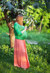 Side view of pregnant woman using digital tablet while standing on grassy field in park