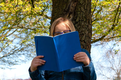 Young school age girl reading from blue textbook in park.