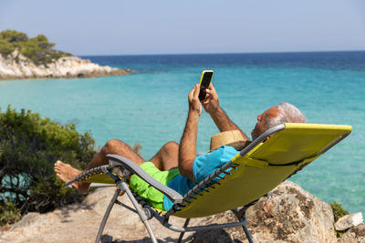 A man on holiday relax in the sun on the deck chair holding his phone chatting with and technology.