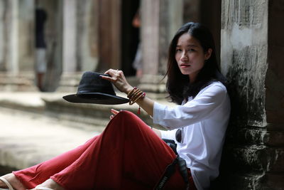 Portrait of young woman holding hat sitting by architectural column at angkor wat