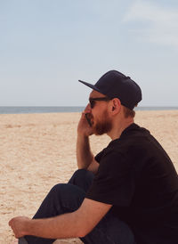 Bearded millennial man in black t-shirt snapback blue seascape, smartphone talking calling friends.