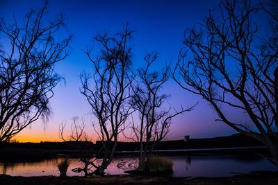 Silhouette bare trees by lake against blue sky during sunset