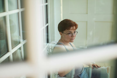 Portrait of young woman looking away while standing against window
