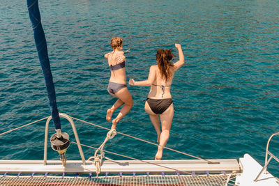 Two women friends jump off catamaran sailing yacht into blue sea in the mediterranean during summer.