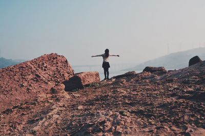 Rear view of woman with arms outstretched standing on rock formation against sky