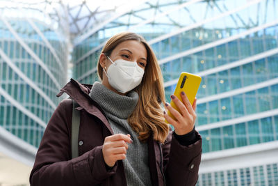 Business woman covering face with protective mask holding mobile phone with modern city background