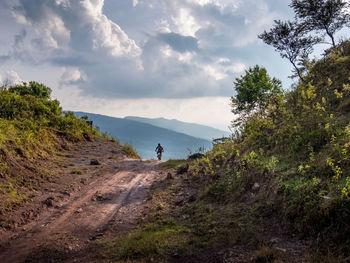 Person riding bicycle on footpath against cloudy sky