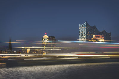 Light trails on illuminated bridge by buildings against sky at night