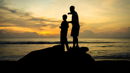 Silhouette father and son standing on rock at beach against sky during sunset