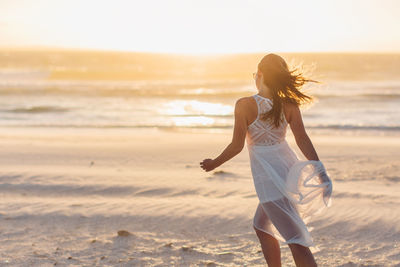 Rear view of young woman walking at beach against clear sky during sunset